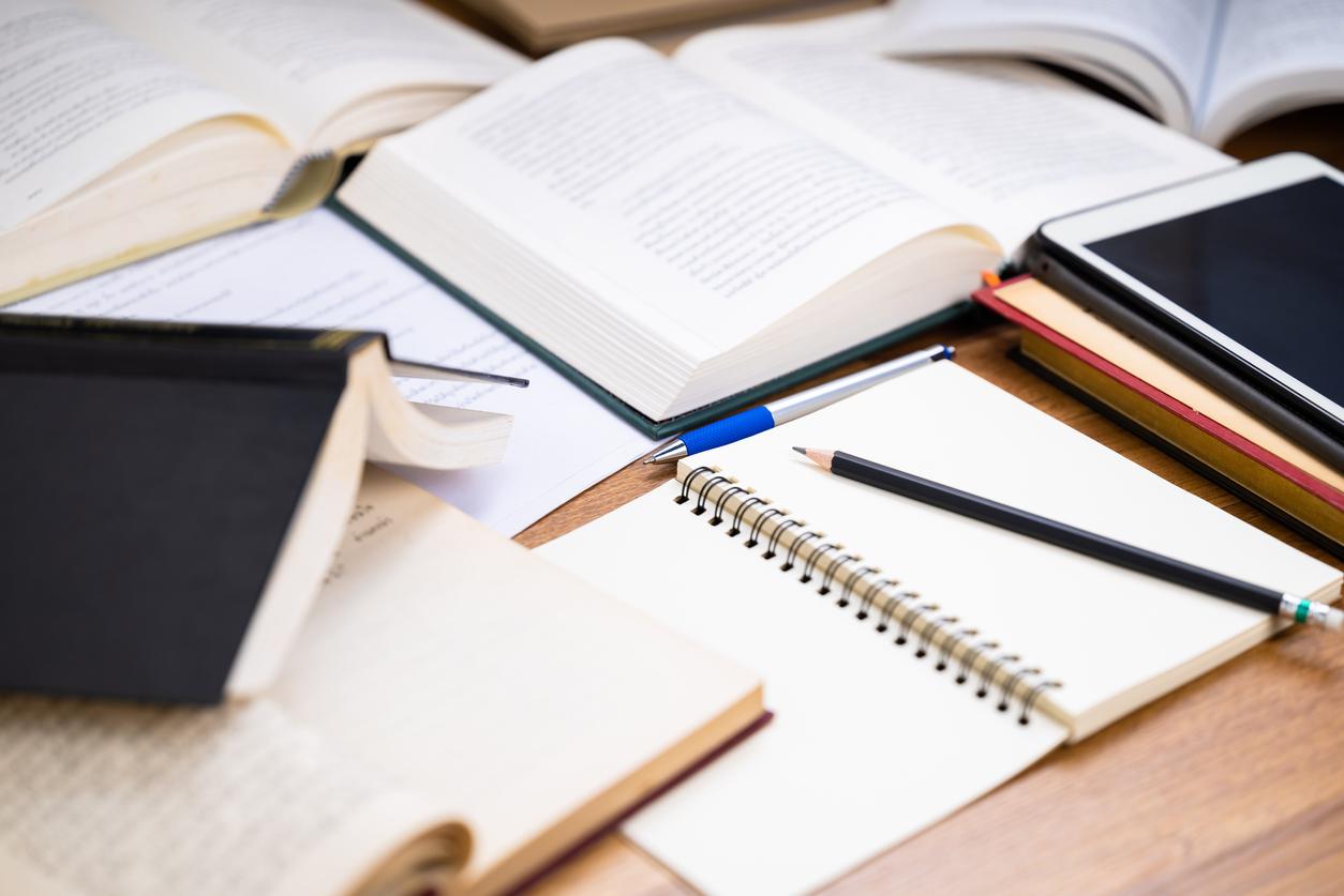 Books and papers piled around an empty notepad with a pencil poised to write